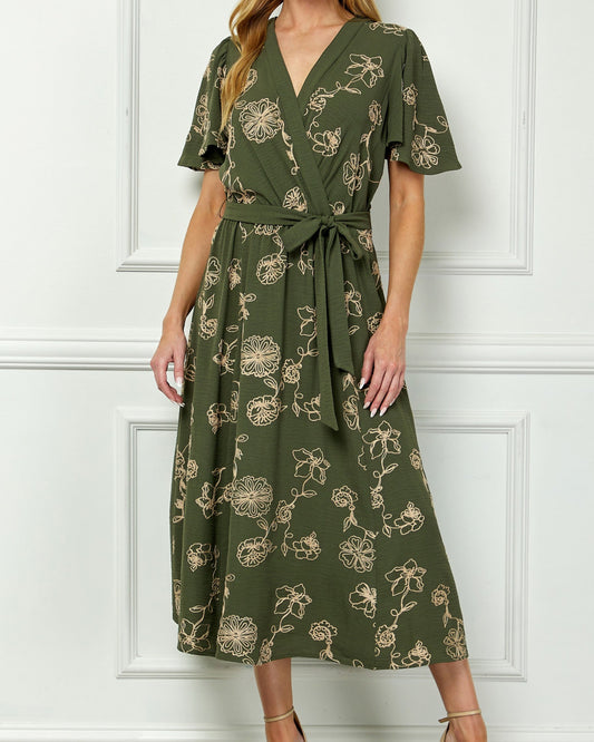 Woman wearing a green floral dress against a white paneled wall.