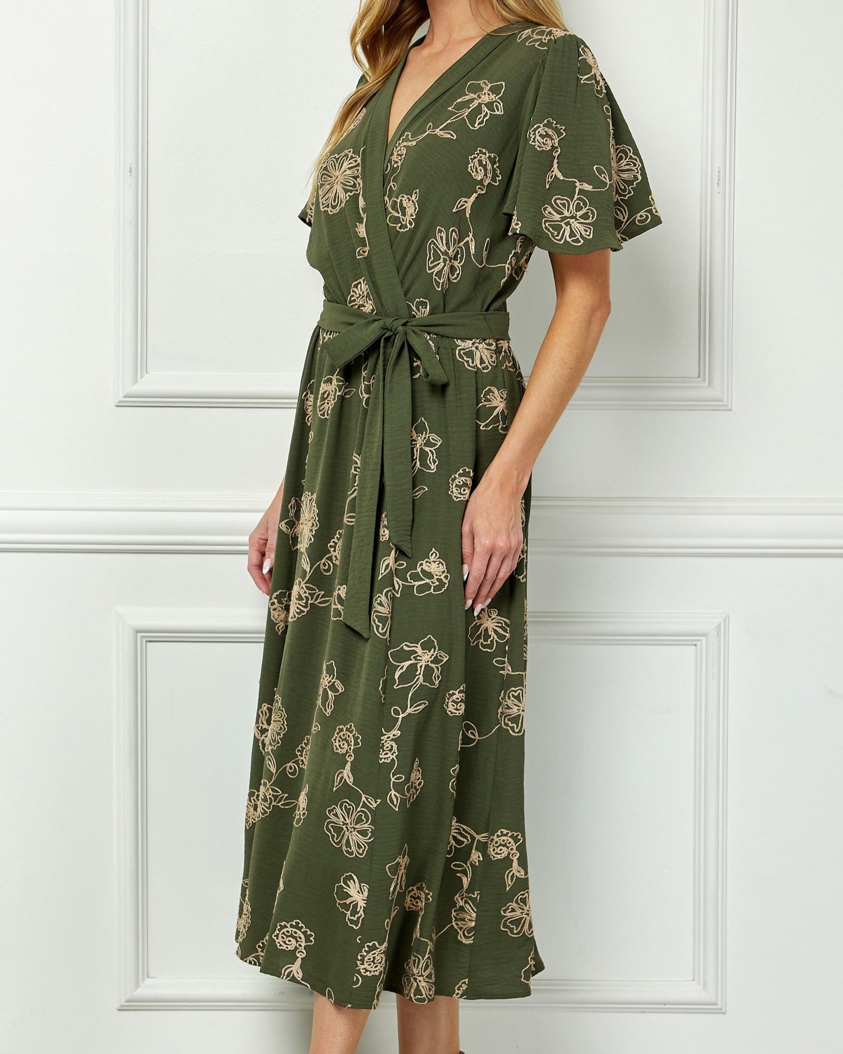 Woman wearing a green floral dress against a white paneled wall.