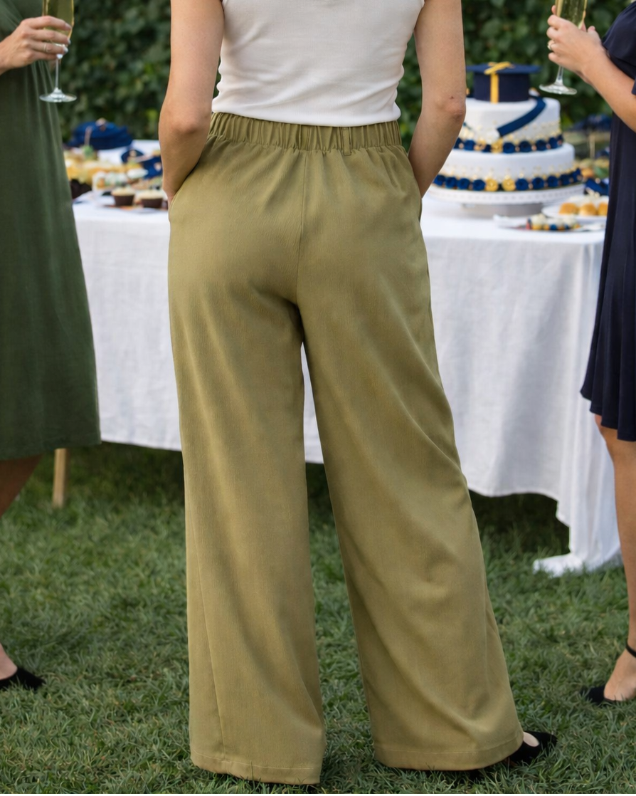 Woman in a white top and green pants standing at a graduation party with decorations and cake.