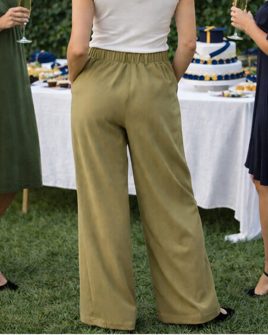 Woman in a white top and green pants standing at a graduation party with decorations and cake.