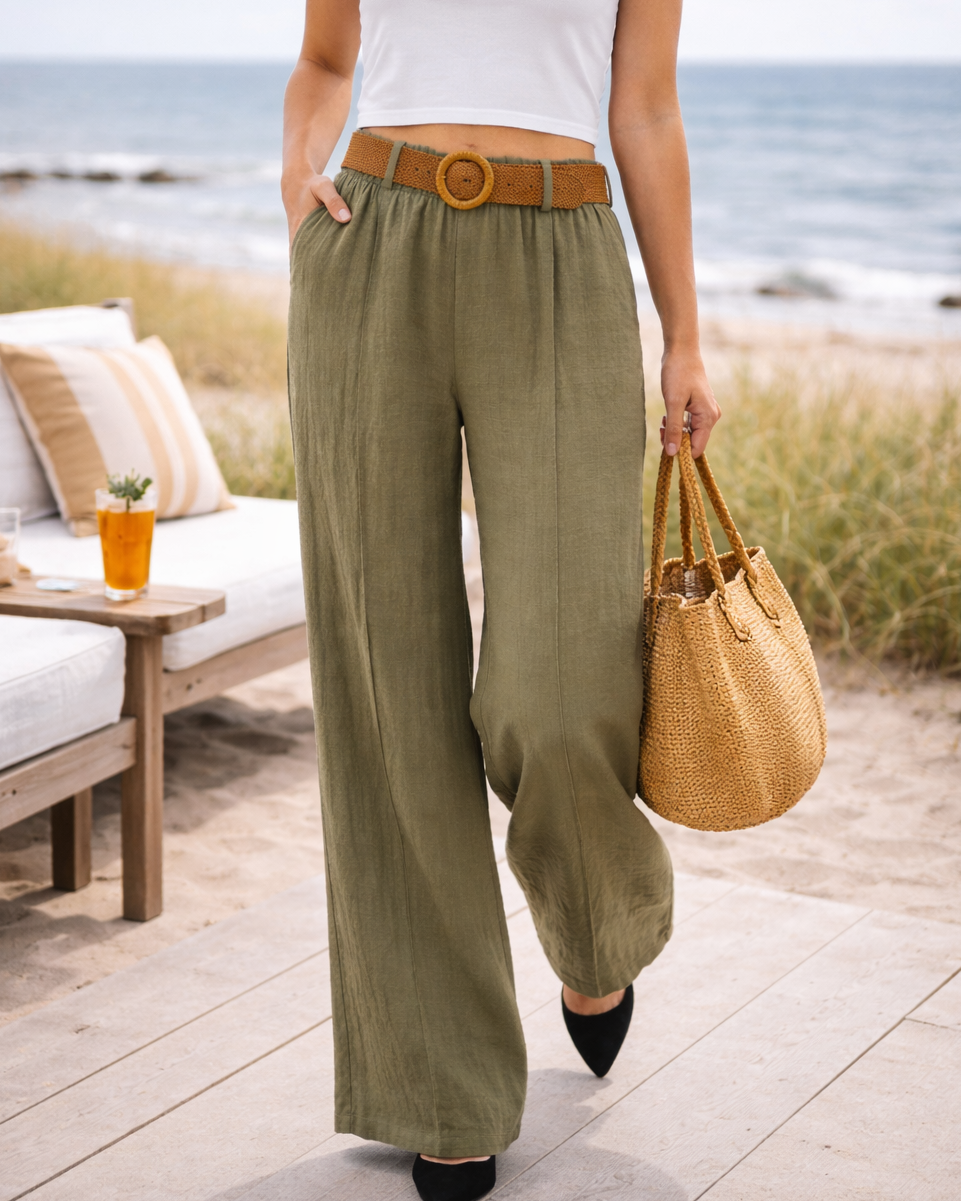 Woman in white top and green pants holding a straw bag on a beach setting.