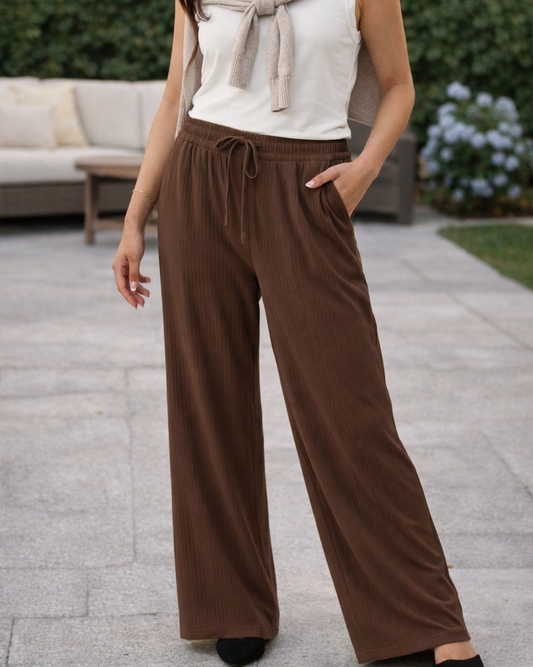 Woman wearing a white top, brown scarf, and wide-leg brown pants standing outdoors.