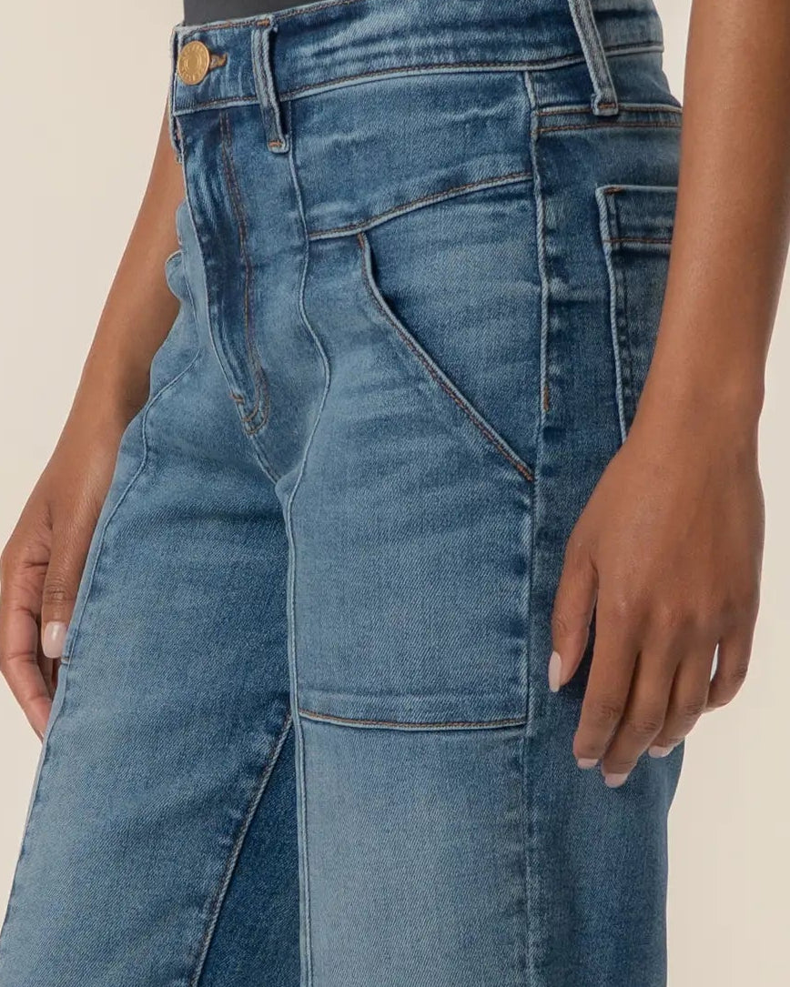 A person wearing medium-wash blue wide-leg jeans with a black top and beige sneakers, shown from the waist down against a plain beige studio background.