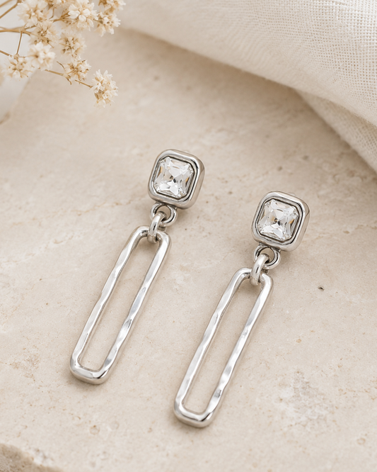 Silver earrings with square gemstones on a beige stone surface with dried flowers.